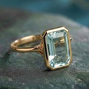 May include: A gold ring featuring a large, rectangular, light blue gemstone. The gemstone is set in a gold bezel, and the ring band is also gold. The ring is displayed on a dark, textured surface, with a blurred blue background.