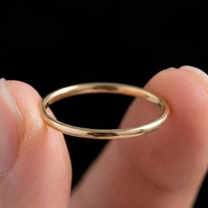 Puede incluir: Un anillo delgado de color dorado con un diseño de banda simple. El anillo se sostiene sobre un fondo negro, resaltando su superficie pulida. El anillo parece ser de estilo minimalista, adecuado para el uso diario.