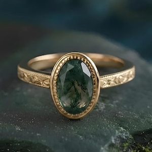 May include: A gold ring with an oval, green gemstone. The gemstone has a unique, moss-like pattern. The ring's band features an intricate, engraved design. The ring is set against a dark, textured background.