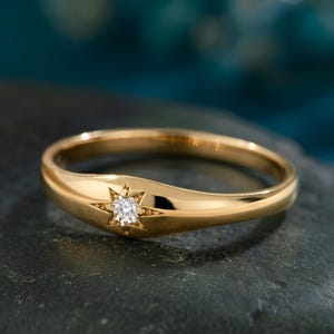 May include: A gold ring with a starburst design and a small, clear gemstone at the center. The ring is set on a dark, textured surface. The ring's band is smooth and polished.