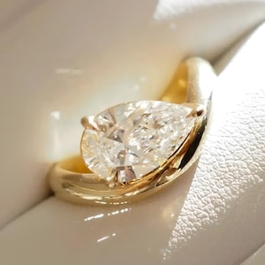 May include: A gold ring with a pear-cut diamond, presented in a white box. The ring features a smooth, curved band that cradles the diamond. The diamond is clear and reflects light.