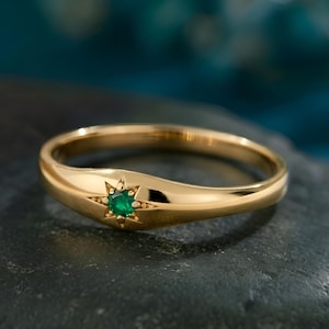 May include: A gold ring with a starburst design and a small, round, green gemstone. The ring is set on a dark, textured surface, with a blurred teal background. The ring's design is eye-catching.