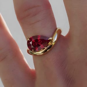 Puede incluir: Un anillo de oro con una piedra preciosa de color rojo intenso en forma de pera, montada en una banda de oro amarillo pulido. La banda se curva alrededor de la base de la piedra, que está asegurada por pequeñas garras. El diseño resalta el contraste entre la gema roja vibrante y el acabado metálico dorado, liso y reflectante.