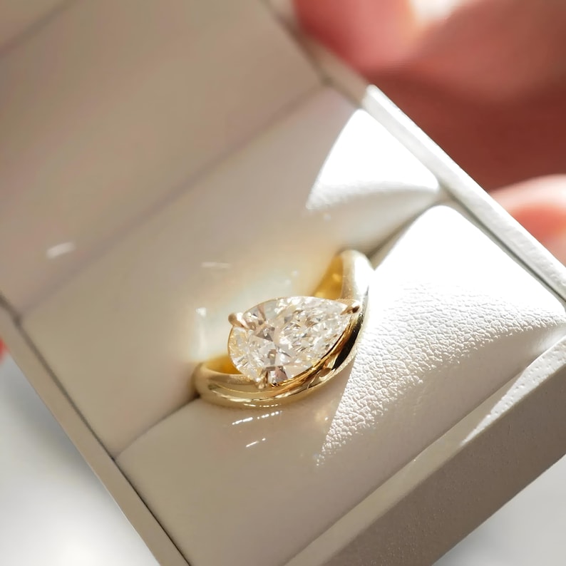 May include: A gold ring with a pear-cut diamond, presented in a white box. The ring features a smooth, curved band that cradles the diamond. The diamond is clear and reflects light.