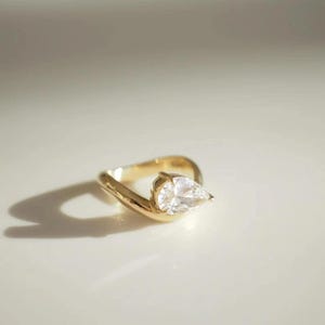 May include: A gold ring with a pear-shaped, clear gemstone. The ring has a unique, curved design, with the gemstone set at an angle. The ring is displayed on a white surface, with a shadow cast to the left.