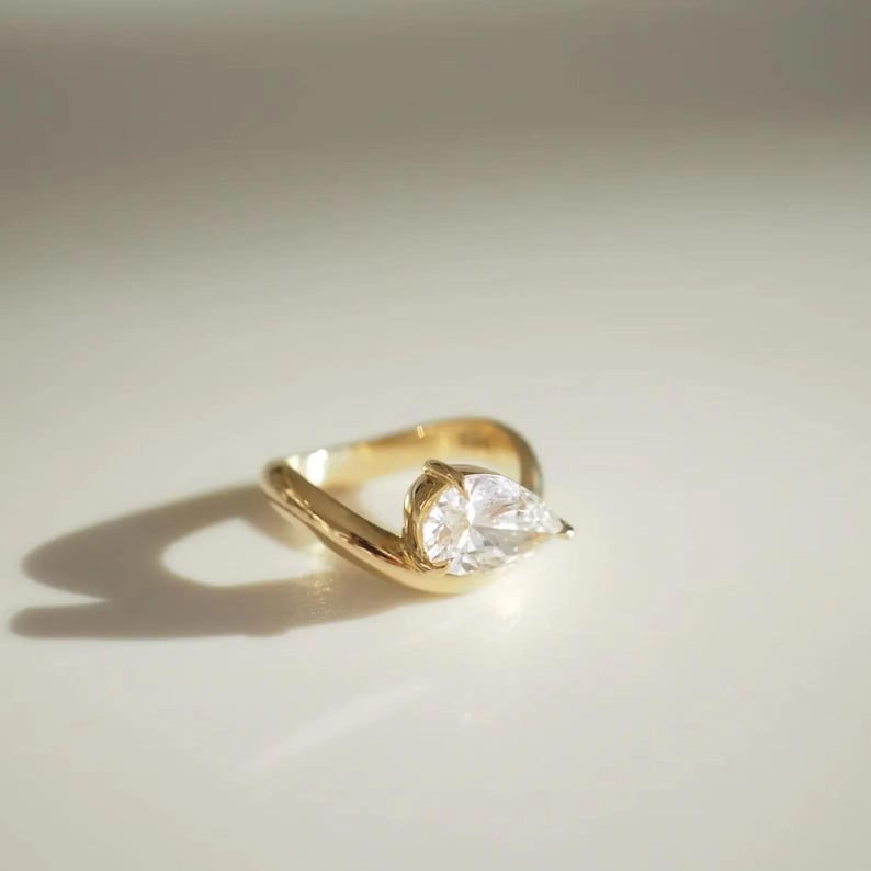 May include: A gold ring with a pear-shaped, clear gemstone. The ring has a unique, curved design, with the gemstone set at an angle. The ring is displayed on a white surface, with a shadow cast to the left.
