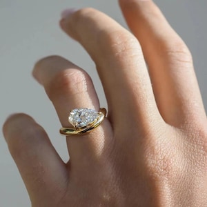 May include: A gold ring with a pear-shaped diamond. The diamond is the focal point, set in a swirling gold band. The ring is shown on a person's finger, catching the light.