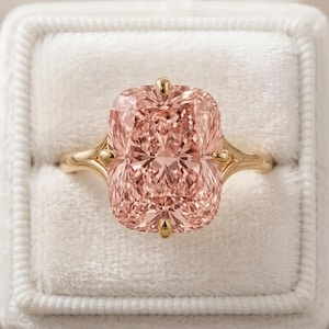 Puede incluir: Un anillo con una gema rosa, engastada en una banda dorada. La piedra de talla cojín es de un tono rosa suave, y la banda dorada presenta detalles delicados. El anillo se presenta en un joyero de terciopelo blanco.