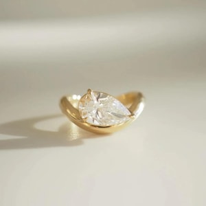May include: A gold ring featuring a pear-shaped, clear gemstone. The ring's band curves around the stone, creating a modern, elegant design. The ring is set against a soft, neutral background, highlighting its brilliance.