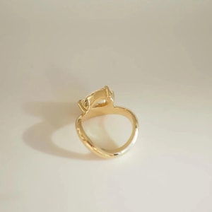 May include: A gold ring with a pear-shaped gemstone. The ring's band is smooth and curves into a rounded shape. The gemstone is clear and faceted, catching the light. The ring is set against a neutral background.