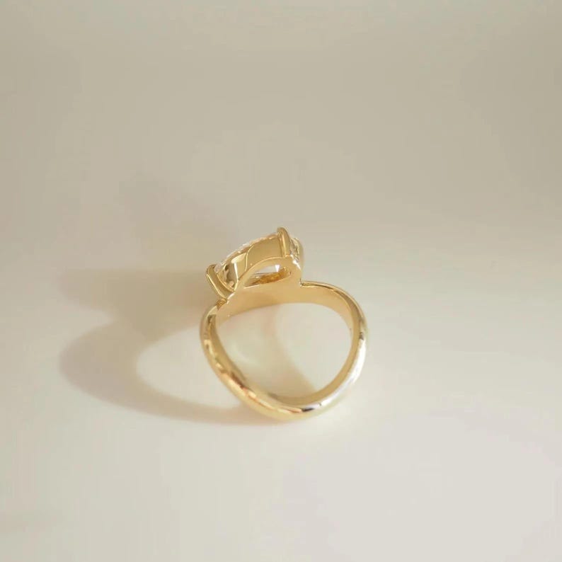 May include: A gold ring with a pear-shaped gemstone. The ring's band is smooth and curves into a rounded shape. The gemstone is clear and faceted, catching the light. The ring is set against a neutral background.