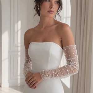 Könnte beinhalten: Ein trägerloses weißes Brautkleid mit langen, transparenten Ärmeln, die mit kleinen, gleichmäßig verteilten Perlen verziert sind. Das Kleid hat ein figurbetontes Oberteil und ein schlichtes, elegantes Design. Die Ärmel verleihen einen Hauch von Raffinesse.