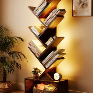 Op de afbeelding: Een donkerbruine, boomvormige boekenplank gevuld met boeken. De boekenplank heeft schuine planken en is van binnenuit verlicht. Een kleine klok en een mand staan op de basis. Een ingelijste foto en een potplant zijn ook zichtbaar.