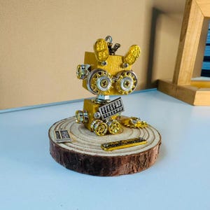 May include: A small robot figurine constructed from yellow painted wooden blocks, metallic gears, and silver-toned hardware. It features two abacus-style charms, one attached to its chest and one resting on the circular wooden base beneath it.