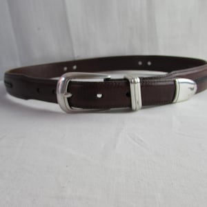 May include: A brown leather belt with a silver buckle and accents. The belt has a classic design with a rectangular buckle and a single belt loop. The leather appears smooth and of good quality.