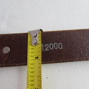 May include: Close-up of a brown leather belt with silver rivets and the number "12000" stamped on it. A yellow measuring tape shows the belt is approximately 2.5 cm wide.