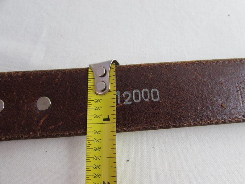 May include: Close-up of a brown leather belt with silver rivets and the number "12000" stamped on it. A yellow measuring tape shows the belt is approximately 2.5 cm wide.