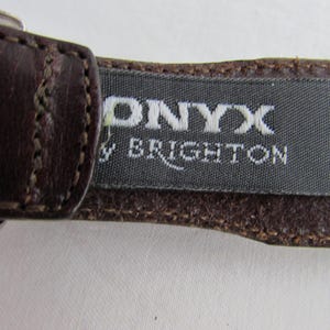 May include: Close-up of a brown leather belt with the brand name "ONYX by BRIGHTON" printed on a grey fabric tag. The belt is stitched with a lighter brown thread.
