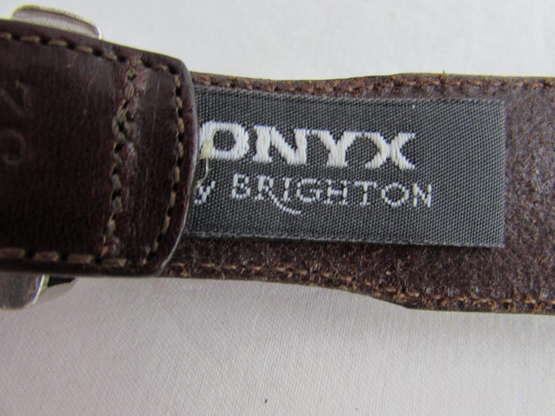 May include: Close-up of a brown leather belt with the brand name "ONYX by BRIGHTON" printed on a grey fabric tag. The belt is stitched with a lighter brown thread.