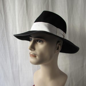 Könnte beinhalten: Ein schwarzer Fedora-Hut mit breiter Krempe und weißem Band. Der Hut wird auf einem Schaufensterkopf vor weißem Hintergrund präsentiert. Das klassische Design des Hutes eignet sich für verschiedene Anlässe.