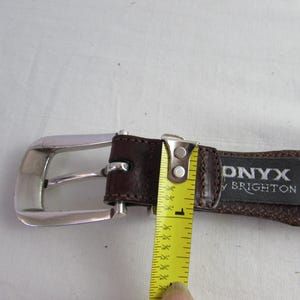 May include: A brown leather belt with a silver buckle. The belt is being measured with a yellow tape measure, showing a width of approximately 2.5 cm. The tag reads "ONYX by BRIGHTON".