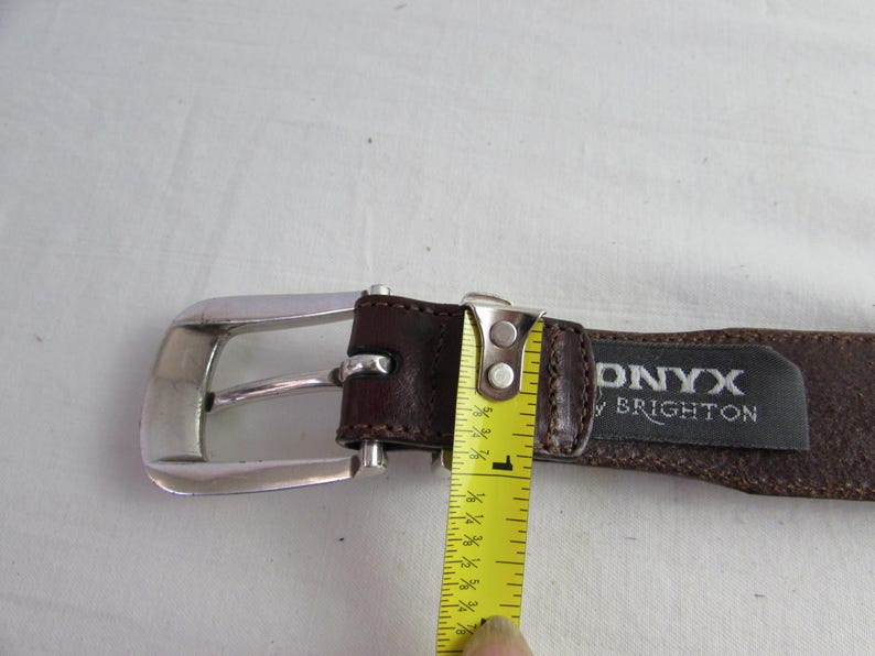 May include: A brown leather belt with a silver buckle. The belt is being measured with a yellow tape measure, showing a width of approximately 2.5 cm. The tag reads "ONYX by BRIGHTON".