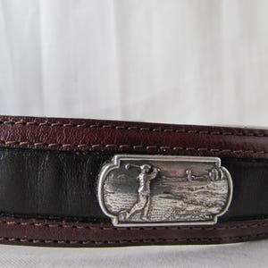 May include: Leather belt with a silver buckle depicting a golfer. The belt has a black central section with burgundy edges and stitching. The buckle is rectangular and shows a detailed golf course scene.