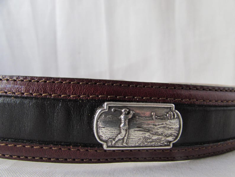 May include: Leather belt with a silver buckle depicting a golfer. The belt has a black central section with burgundy edges and stitching. The buckle is rectangular and shows a detailed golf course scene.