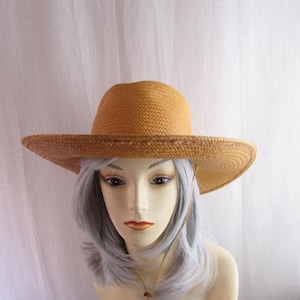 May include: A straw hat with a wide brim and a tan color. The hat has a rounded crown and a chin strap. The hat is displayed on a mannequin head with gray hair.