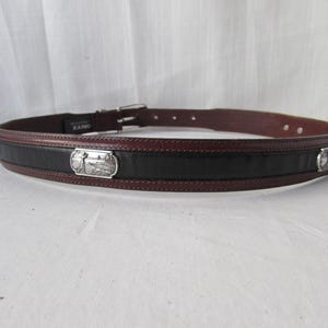 May include: A brown and black leather belt with a silver buckle. The belt has a black centre section and brown edges. Silver rectangular plaques with a landscape design are on the front of the belt.