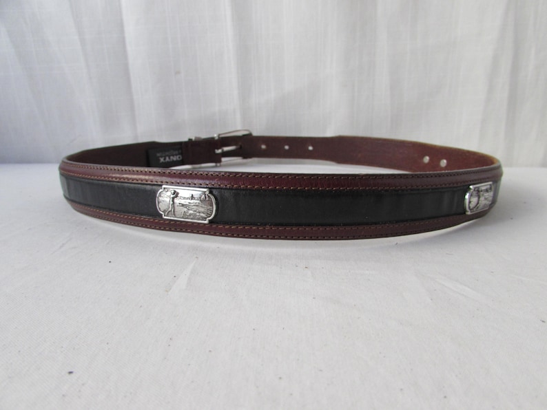 May include: A brown and black leather belt with a silver buckle. The belt has a black centre section and brown edges. Silver rectangular plaques with a landscape design are on the front of the belt.