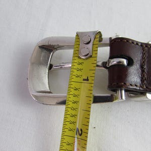 May include: Close-up of a brown leather belt with a silver buckle. The belt is measured with a yellow tape measure, showing a size of 81 cm. The belt is on a white surface.
