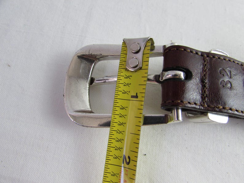 May include: Close-up of a brown leather belt with a silver buckle. The belt is measured with a yellow tape measure, showing a size of 81 cm. The belt is on a white surface.