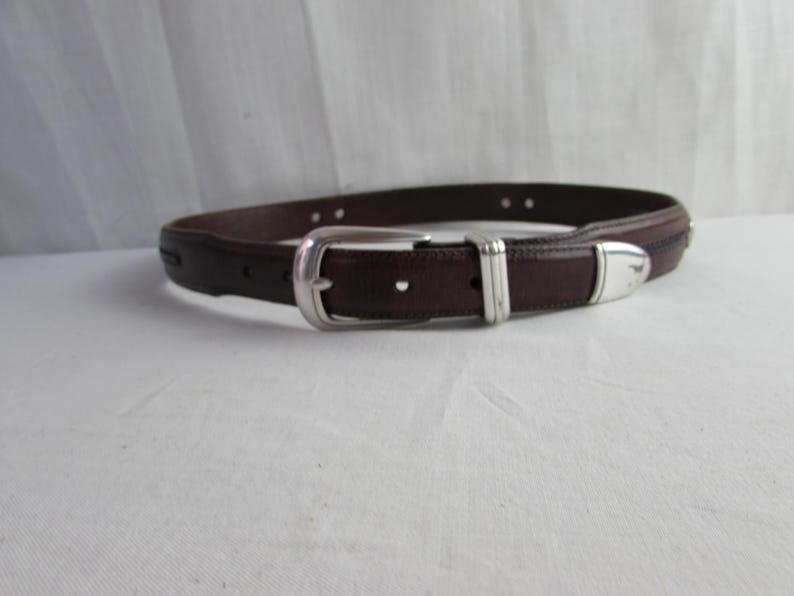 May include: A brown leather belt with a silver buckle and accents. The belt has a classic design with a rectangular buckle and a single belt loop. The leather appears smooth and of good quality.