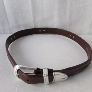 May include: A brown leather belt with a silver buckle and accents. The belt has a classic design with a rectangular buckle and a decorative silver piece near the end. The belt is a medium width and has several holes for adjustment.
