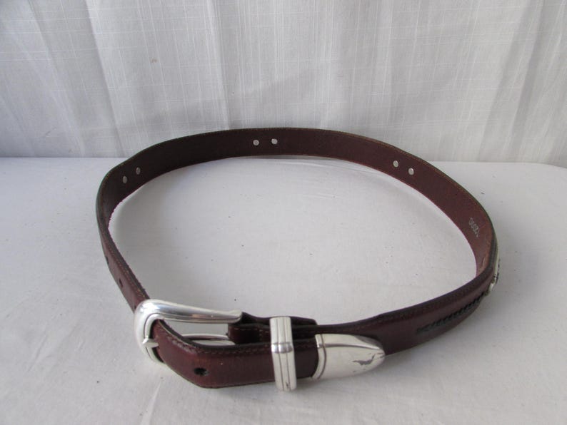 May include: A brown leather belt with a silver buckle and accents. The belt has a classic design with a rectangular buckle and a decorative silver piece near the end. The belt is a medium width and has several holes for adjustment.
