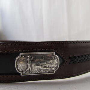 May include: A brown leather belt with a silver rectangular buckle depicting a golfer. The belt features a black central section and a braided leather detail. The belt is made of leather.