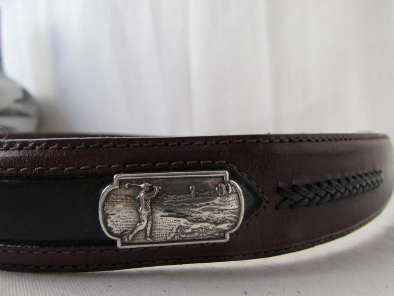 May include: A brown leather belt with a silver rectangular buckle depicting a golfer. The belt features a black central section and a braided leather detail. The belt is made of leather.
