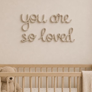 May include: A wall-mounted sign featuring the beige rope-textured cursive text "you are so loved" displayed in two lines. The letters are formed from a thick, twisted cord material, creating a soft, tactile appearance against the wall surface.