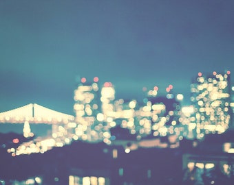 May include: Nighttime cityscape with blurred lights. A bridge with illuminated lights is in the foreground, with tall buildings in the background. The lights are a mix of white, yellow, and red against a dark blue sky.