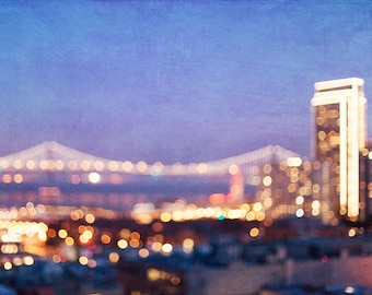 May include: A blurred image of a city skyline at night with a bridge in the background. The city lights are out of focus and create a soft, warm glow. The sky is a deep blue color.