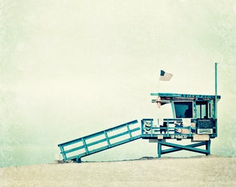 May include: A blue lifeguard stand with a ramp leading up to it. The stand has a sign that reads "Adina For L." and an American flag is flying from the top. The stand is on a sandy beach with a light blue sky in the background.