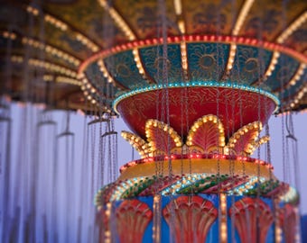 May include: Close-up of a brightly lit carousel ride. The ride features a red, teal, yellow, and blue color scheme with decorative lights. The ride's canopy is adorned with lights, and chains hang down, ready for riders.