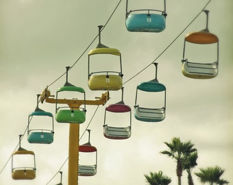 May include: A colorful aerial tramway with multiple cabins, each with a different number. The cabins are suspended from cables against a cloudy sky. Palm trees are visible in the background.