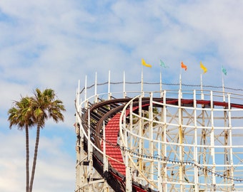 May include: A white wooden roller coaster track with a red wooden track running through it. The roller coaster is against a blue sky with white clouds. There are five colorful flags at the top of the roller coaster.