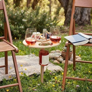 Pode incluir: Uma mesa de vinho redonda de madeira para exterior com um suporte central para garrafas e duas ranhuras laterais para copos de vinho. A mesa contém uma garrafa de vinho tinto e dois copos cheios de vinho tinto, junto a um prato com figos e queijo.