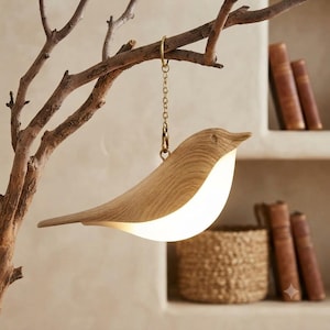 May include: A bird-shaped hanging lamp featuring a light-colored wood grain body with a smooth, curved silhouette. The lamp emits a soft, warm glow from its underside and is suspended by a thin gold-toned metal chain from a decorative branch.