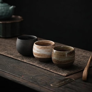 May include: Three small ceramic tea cups arranged in a row. The first cup is matte black, the second features a white and brown marbled glaze, and the third has a mottled green and brown finish. Each cup has a rounded, handleless bowl shape.