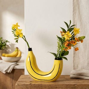 Puede incluir: Un jarrón en forma de plátano amarillo con rayas negras, que contiene flores amarillas y naranjas. El jarrón está colocado sobre una superficie de madera, con un cuenco de plátanos y una planta en el fondo. Un artículo de decoración del hogar único.