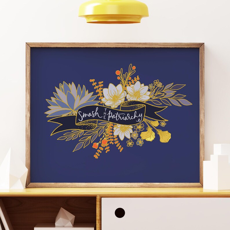 May include: A framed art print with a navy blue background. The artwork features a floral arrangement in gold, white, and orange hues, with a banner that reads "Smash the Patriarchy" in white script. The frame is a warm brown color.